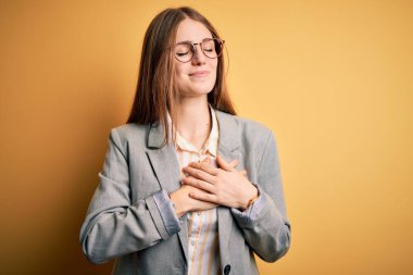 Sarı arka planda gözlüklü, ceketli ve gözlüklü genç, güzel bir kadın. Elleri göğsünde, gözleri kapalı ve yüzünde minnet dolu bir jest ile gülümsüyor. Sağlık kavramı.