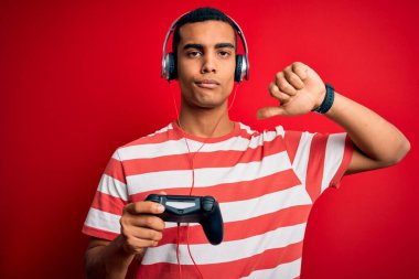 Yakışıklı Afrikalı Amerikalı oyuncu, video oyunu oynuyor. Jostick ve kızgın yüzlü kulaklıklar kullanıyor. Negatif işaretler, baş parmakları aşağıda, reddedilme kavramı.