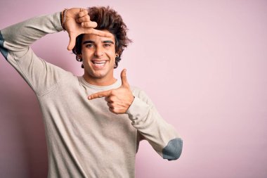 İzole edilmiş pembe arka plan üzerinde sıradan tişörtler giyen genç yakışıklı adam gülümsüyor ve elleri ve parmakları mutlu yüzlü bir çerçeve yapıyor. Yaratıcılık ve fotoğrafçılık kavramı.