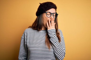 Genç, güzel esmer kadın Fransız beresi takıyor ve sarı arka planda gözlük takıyor. Elle ağzını kapatmaktan sıkılmış. Huzursuz ve uykulu..