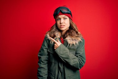 Kayak gözlüğü takan ve kırmızı arka planda kayak için kışlık ceket giyen genç sarışın kız endişeli, endişeli ve şaşırmış bir ifade takınıyor.