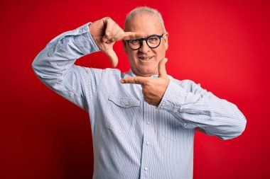 Orta yaşlı yakışıklı, beyaz, sıradan çizgili gömlek ve kırmızı arka planda gözlük takan, gülümseyen, elleri ve parmakları mutlu yüzlü bir çerçeve yapan adam. Yaratıcılık ve fotoğrafçılık kavramı.