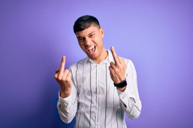 Young handsome hispanic man wearing elegant business shirt standing over purple background Showing middle finger doing fuck you bad expression, provocation and rude attitude. Screaming excited