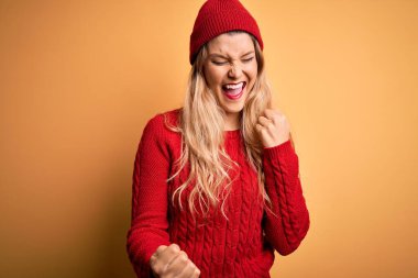 Beyaz arka plan üzerine günlük kazak ve yün şapka giyen genç, güzel sarışın kadın kolları havada ve gözleri kapalı olarak başarıyı kutluyor ve şaşırıyor. Kazanan konsept.