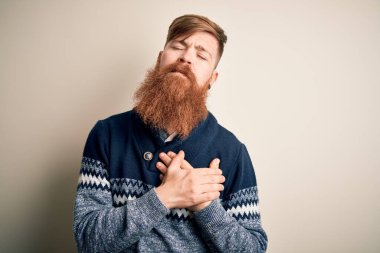 Sarışın, sakallı, kışın süveter giyen, izole edilmiş arka planda gülümseyen elleri göğsünde ve yüzünde minnet ifadesi olan yakışıklı bir İrlandalı. Sağlık kavramı.