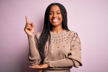 Pembe izole edilmiş arka planda moda süveteri giyen Afro-Amerikalı genç bir kadın yüzünde kocaman bir gülümsemeyle kameraya el ve parmak ile bakıyor..