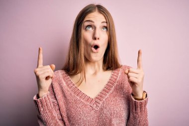 Genç, güzel, kızıl saçlı, günlük süveter giyen bir kadın pembe arka planda şaşkın şaşkın yukarı bakıyor ve parmaklarıyla işaret ediyor ve kollarını kaldırıyordu..