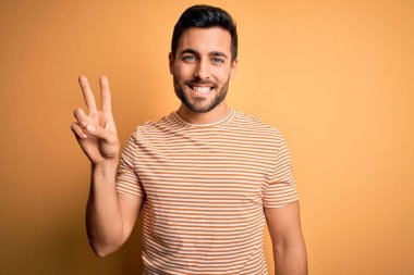 Sakallı, sarı arka planda günlük çizgili tişört giyen genç ve yakışıklı bir adam iki numarayı gösterirken kendinden emin ve mutlu bir şekilde gülümsüyor..