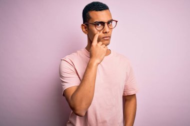 Yakışıklı Afro-Amerikan erkeği pembe arka planda günlük tişört ve gözlük takıyor. Gözünü dikip jestini izliyor. Şüpheli bir ifade.