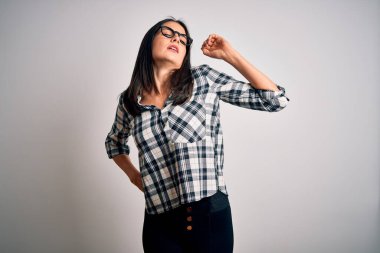 Mavi gözlü genç esmer kadın, günlük gömlek ve gözlük takıyor beyaz arka planda geriniyor, yorgun ve rahat, uykulu ve sabahın erken saatlerinde esniyor.