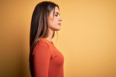 Genç, güzel, esmer bir kadın, günlük tişört giyiyor sarı arka planda duruyor, yan tarafa bakıyor, rahat bir görünüm takınıyor ve kendine güvenen bir gülümseme sergiliyor..