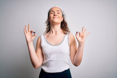 Kıvırcık saçlı güzel bir sporcu kadın izole edilmiş beyaz arka planda spor giyer rahatlar ve parmaklarıyla meditasyon hareketi yaparken gözleri kapalı gülümser. Yoga konsepti.