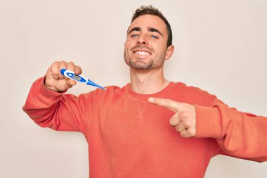 Mavi gözlü, dijital termometre kullanarak sıcaklığı ölçen genç yakışıklı bir adam mutlu bir gülümsemeyle eli ve parmağı ile işaret ediyor.