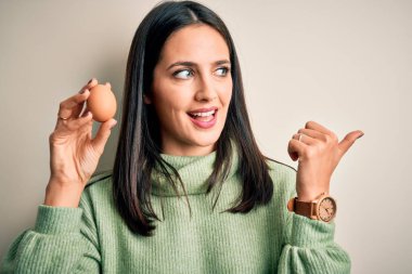 Mavi gözlü genç esmer kadın izole edilmiş arka planda taze çiğ yumurta tutuyor ve gülümseyen yüzüyle baş parmağıyla işaret ediyor.