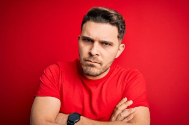 Kırmızı izole edilmiş arka plan üzerine sıradan tişört giyen mavi gözlü genç adam şüpheci ve gergin, yüzünde çapraz kollu tasvip etmeyen bir ifade var. Negatif kişi.