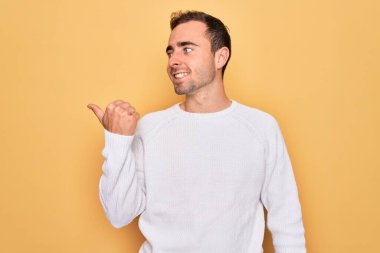 Mavi gözlü, sıradan bir süveter giyen, sarı arka planda duran mutlu yüzüyle gülümseyen ve baş parmağıyla yan tarafı gösteren genç yakışıklı bir adam..