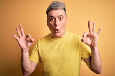 Sarı izole edilmiş arka plan üzerinde sarı tişört giyen genç, yakışıklı ve modern bir adam parmaklarıyla onay sembolü yaparken şaşırmış ve şok olmuş görünüyor. Çılgın ifade