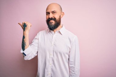 Sakallı, yakışıklı, zarif gömlekli, pembe arka planda mutlu yüzüyle gülümseyen ve baş parmağıyla yüzünü gösteren kel bir adam..
