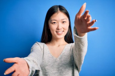 Genç ve güzel Asyalı kadın, günlük süveter giyip mavi izole edilmiş arka planda dikiliyor ve kameraya sarılıp gülümsüyor. Mutluluğu kucaklayan neşeli bir ifade..