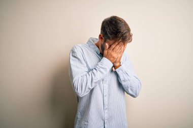 Beyaz arka planda duran çizgili tişörtlü, sakallı, genç ve yakışıklı bir adam. Ağlarken yüzünü ellerle kapatan üzgün bir ifade. Depresyon kavramı.