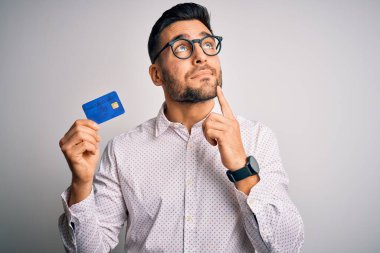Young business man holding credit card over isolated background serious face thinking about question, very confused idea