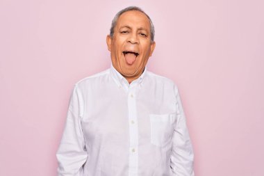 Üst düzey yakışıklı, gri saçlı, zarif tişörtlü, soyutlanmış pembe arka planda, komik bir ifadeyle dilini dışarı çıkaran adam. Duygu kavramı.