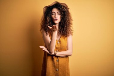 Güzel esmer kadın tatilde. Kıvırcık saçlı, piercing takan, şapkalı, elbisesi olan, kameraya bakıp havada öpüşen güzel ve seksi bir kadın. Aşk ifadesi.