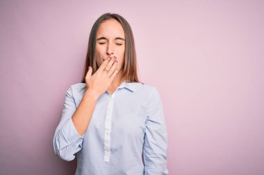 Zarif gömlekler giyen genç ve güzel iş kadını izole edilmiş pembe arka planda dikiliyor. Elle ağzını kapatmaktan sıkılmış. Huzursuz ve uykulu..