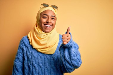 Sarı başörtüsü takan ve güneş gözlüğü takan genç Afrikalı Amerikalı öğrenci el hareketleriyle mutlu mesut başparmak hareketi yapıyor. Kameraya bakıp başarıyı gösteren ifade onaylanıyor.