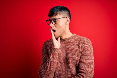 Kırmızı arka planda inek gözlüğü takan genç, yakışıklı İspanyol adam ağzıyla ağzını kapatmaktan sıkılmış. Huzursuz ve uykulu..