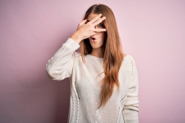 Genç, güzel, kızıl saçlı bir kadın pembe arka plan üzerinde gözlük ve süveter takıyor. Şok olmuş bir şekilde yüzünü ve gözlerini kapıyor. Utanmış bir ifadeyle parmaklarının arasından bakıyor..