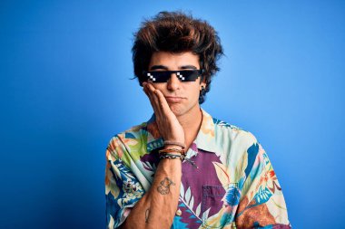 Gangster gözlüğü takan genç yakışıklı adam mavi arka planda duruyor. Kollarını kavuşturup depresyondan sıkılmış ve yorgun görünüyor..