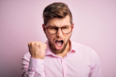 Sakallı, mavi gözlü, pembe gömlekli, gözlüklü kızgın ve kızgın genç, sarışın bir adam öfkeyle bağırırken yumruğunu kaldırıyor. Öfke ve agresif kavram.