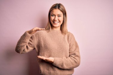 Pembe, izole edilmiş arka planda yün süveter giyen genç, güzel sarışın kadın. Ellerinde büyük ve büyük beden işareti, ölçü sembolü var. Kameraya bakıp gülümsüyor. Ölçüm konsepti.