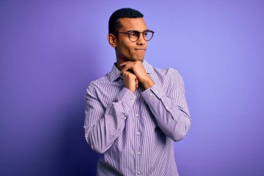 Yakışıklı Afro-Amerikalı adam mor arka planda çizgili gömlek ve gözlük takıyor. Gergin ve heyecanlı bir şekilde çenesinin kenarına bakıyor.