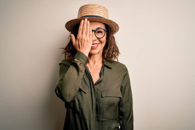 Orta yaşlı esmer kadın gözlük takıyor ve şapka takıyor. İzole edilmiş beyaz arka planda duruyor. Bir gözünü eliyle kapatıyor..