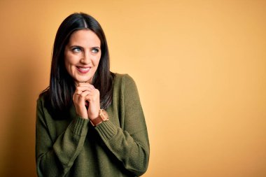 Sarı arka planda yeşil süveter giyen, mavi gözlü genç esmer kadın. Gergin ve heyecanlı bir şekilde çenesinin kenarına bakıyor.