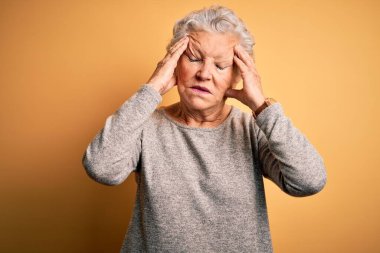 Son sınıf güzeli bir kadın günlük tişört giyip izole edilmiş sarı arka planda dikiliyor. Ağrı ve migrenden dolayı baş ağrısı ve stresten muzdarip. Ellerini başının üstüne koy..