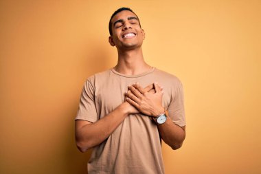 Sarı arka plan üzerinde sıradan tişörtler giyen genç, yakışıklı Afro-Amerikalı adam elleri göğsünde, gözleri kapalı ve yüzünde minnet dolu bir jest ile gülümsüyor. Sağlık kavramı.