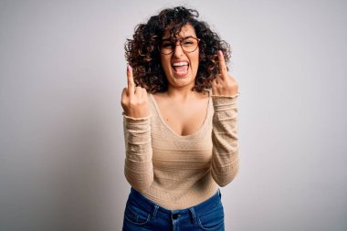 Young beautiful curly arab woman wearing casual t-shirt and glasses over white background Showing middle finger doing fuck you bad expression, provocation and rude attitude. Screaming excited