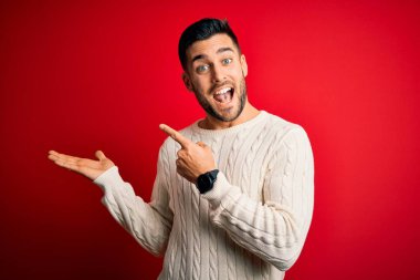 İzole edilmiş kırmızı arka plan üzerinde duran gündelik beyaz süveter giyen genç yakışıklı adam hayrete düşmüş ve kameraya gülümserken eliyle ve parmağıyla işaret ediyordu..