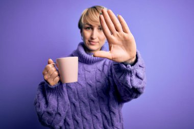 Kısa saçlı, kısa saçlı, kışlık kazak giyen, eli açık bir fincan kahve içen, ciddi ve kendinden emin bir ifadeyle dur işareti yapan genç sarışın kadın, savunma jesti.