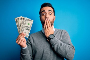 Sakallı ve sakallı yakışıklı bir adam mavi arka planda bir deste banknot tutuyor ağzı kapalı hata yaptığı için utancından ağzı açık, korku ifadesi, sessizlikten korkmak, gizli konsept