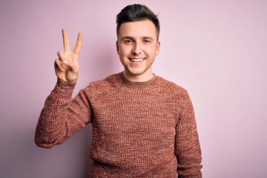 Pembe izole edilmiş arka plan üzerine sıradan bir kazak giyen yakışıklı beyaz bir adam iki numarayı gösterirken kendinden emin ve mutlu bir şekilde gülümsüyor..