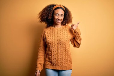 Afro saçlı, sarı arka planda günlük süveter giyen, gülümseyen ve baş parmağıyla yüzünü gösteren Afro Amerikalı genç bir kadın..