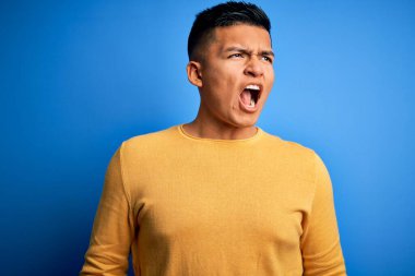İzole edilmiş mavi arka planda sarı süveter giyen genç ve yakışıklı Latin adam sinirli ve öfkeli çığlıklar atıyor, öfkeyle bağırıyor. Öfke ve agresif kavram.