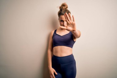 Beyaz arka plan üzerine spor giyerek spor yapan genç ve güzel sarışın spor kadın avuç içiyle şarkı söylemeyi kesiyor. Yüzünde olumsuz ve ciddi bir jest olan uyarı ifadesi.