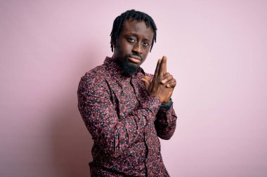 Genç, yakışıklı, Afro-Amerikalı bir adam pembe arka plan üzerinde dikilmiş, el hareketleriyle sembolik bir silah tutuyor, cinayet silahlarıyla oynuyor, kızgın surat ifadesi takınıyor.