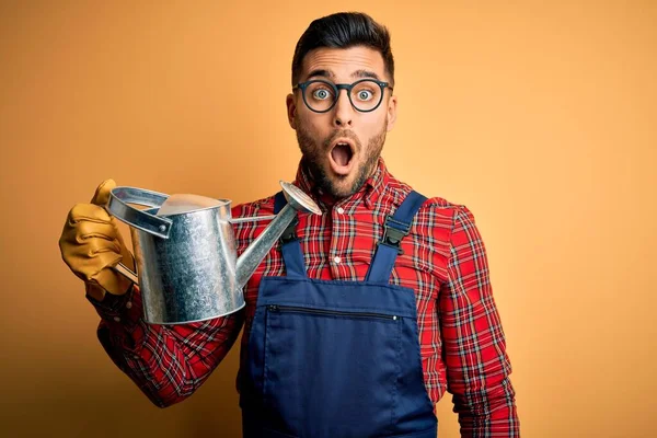 Young gardener man wearing working apron and gloves holding watering can scared in shock with a surprise face, afraid and excited with fear expression