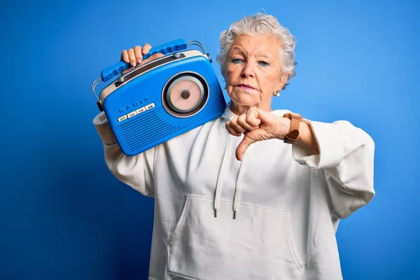 Senior beautiful woman holding vintage radio standing over isolated blue background with angry face, negative sign showing dislike with thumbs down, rejection concept